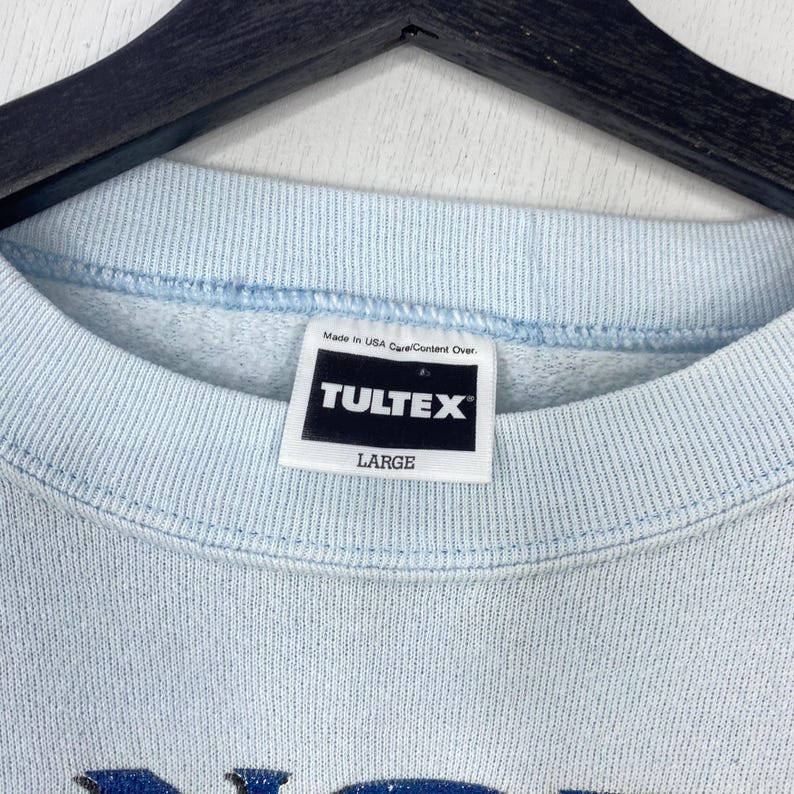May include: A light blue crewneck sweatshirt featuring a white interior tag with black text that reads TULTEX and LARGE. The garment is displayed on a black hanger, showing the ribbed collar and the brand label positioned at the center neckline.
