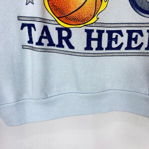 May include: A light blue sweatshirt featuring a graphic of a basketball with yellow flames and the text TAR HEELS in dark blue letters. The design includes a silver horizontal banner and small stars, with a ribbed hem at the bottom of the garment.