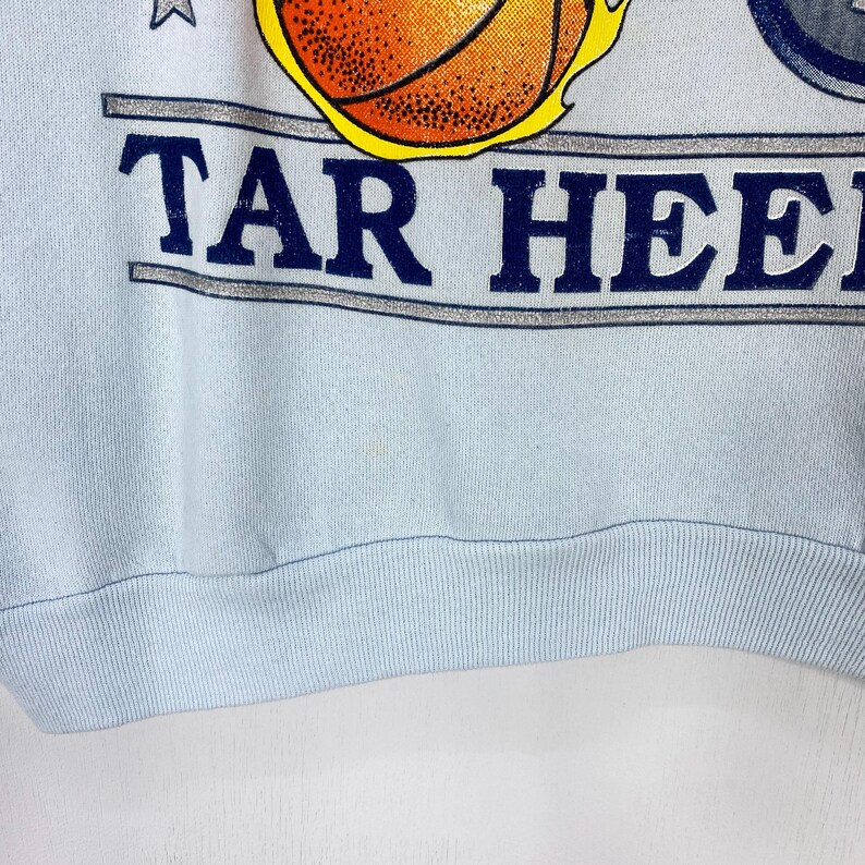 May include: A light blue sweatshirt featuring a graphic of a basketball with yellow flames and the text TAR HEELS in dark blue letters. The design includes a silver horizontal banner and small stars, with a ribbed hem at the bottom of the garment.
