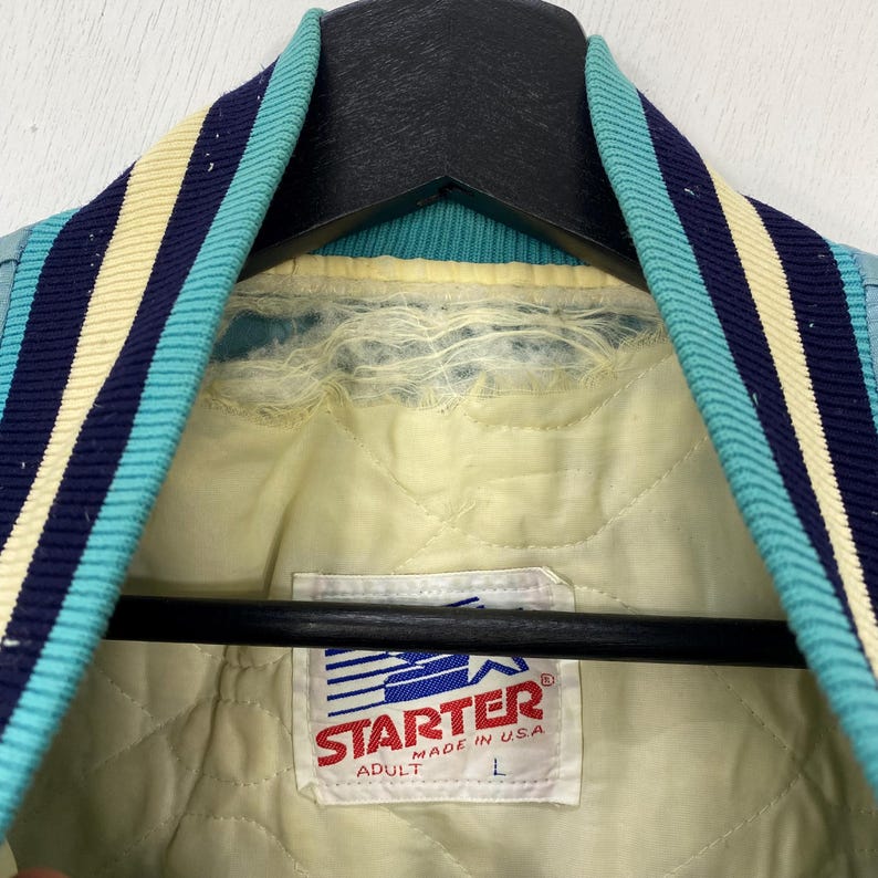 May include: Close-up of a vintage Starter jacket's interior. The jacket features a light blue and navy blue striped collar and a yellow quilted lining. The Starter label is visible, indicating it's an adult size L, made in the USA.