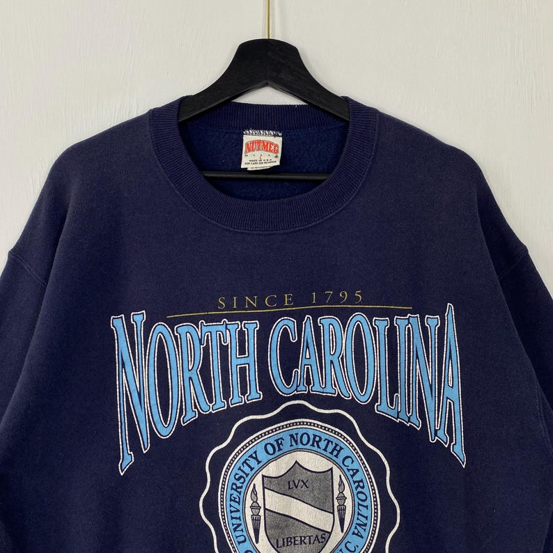 K&ouml;nnte beinhalten: Marineblaues Sweatshirt mit dem Schriftzug "NORTH CAROLINA" in hellblauer, umrandeter Schrift. Das Sweatshirt zeigt auch ein Universit&auml;tswappen und den Text "SINCE 1795". Das Markenetikett "NUTMEG" ist am Ausschnitt sichtbar.
