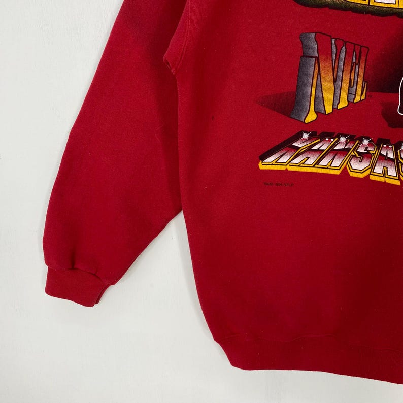 May include: A red long-sleeve sweatshirt featuring a graphic print with the text NFL and KANSAS CITY in a bold, three-dimensional yellow and white font. The garment has ribbed cuffs and a crew neckline, showcasing a vibrant red fabric throughout.