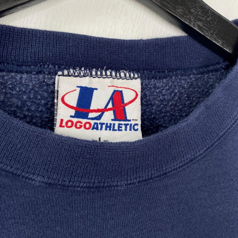 May include: Close-up of a navy blue sweatshirt with the "LA LOGOATHLETIC" tag visible. The tag features a red and blue logo. The sweatshirt is made of a soft, textured material.
