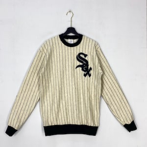 Op de afbeelding: Een crèmekleurig sweatshirt met lange mouwen, voorzien van dunne verticale zwarte krijtstrepen, een zwarte ronde hals en zwarte geribbelde boorden bij de mouwen en zoom. Een groot zwart-wit geborduurd Chicago White Sox logo op de borst.