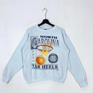 May include: A light blue crewneck sweatshirt featuring a graphic of a basketball going through a flaming hoop. The text reads NORTH CAROLINA TAR HEELS. The design includes four stars and a circular logo with the letters NC in a dark blue font.