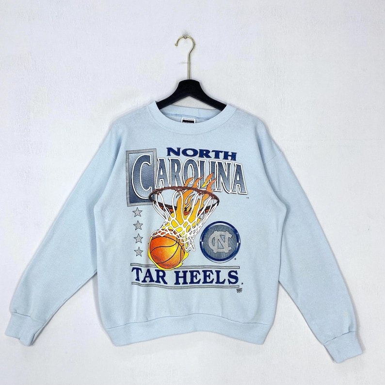 May include: A light blue crewneck sweatshirt featuring a graphic of a basketball going through a flaming hoop. The text reads NORTH CAROLINA TAR HEELS. The design includes four stars and a circular logo with the letters NC in a dark blue font.