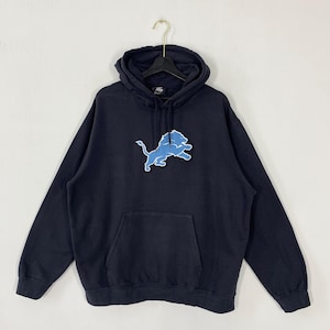 May include: Navy blue hooded sweatshirt with a light blue and white Detroit Lions logo on the front. The sweatshirt has a kangaroo pocket and a drawstring hood. The garment is hanging on a black hanger.