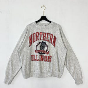 Könnte beinhalten: Ein meliertes graues Sweatshirt mit dem Schriftzug "NORTHERN ILLINOIS" in roten, gebogenen Buchstaben. Ein kreisförmiges Emblem mit einem Husky-Hund befindet sich unter dem Wort "NORTHERN". Das Sweatshirt hängt an einem schwarzen Kleiderbügel.