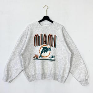 Puede incluir: Una sudadera gris claro con la palabra "MIAMI" en letras de bloque naranja y verde. Debajo del texto hay un gráfico de un delfín saltando a través de un círculo, con la letra "M" dentro. La sudadera está colgada en una percha negra.