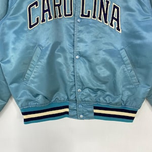 May include: Light blue satin jacket with the text "CAROLINA" in white and navy blue. The jacket has a ribbed hem and cuffs with navy blue and white stripes. Features a button-up front and two front pockets.