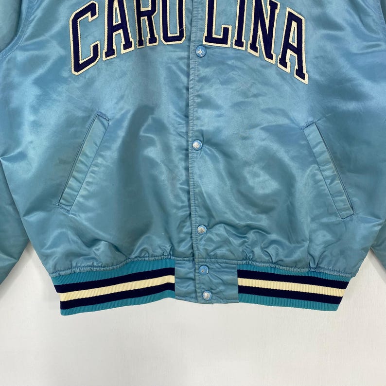 May include: Light blue satin jacket with the text "CAROLINA" in white and navy blue. The jacket has a ribbed hem and cuffs with navy blue and white stripes. Features a button-up front and two front pockets.