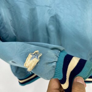 May include: Light blue jacket sleeve with a star patch. The sleeve has a ribbed cuff with stripes in teal, navy, and cream. The star patch is embroidered with a light blue star on a tan background.