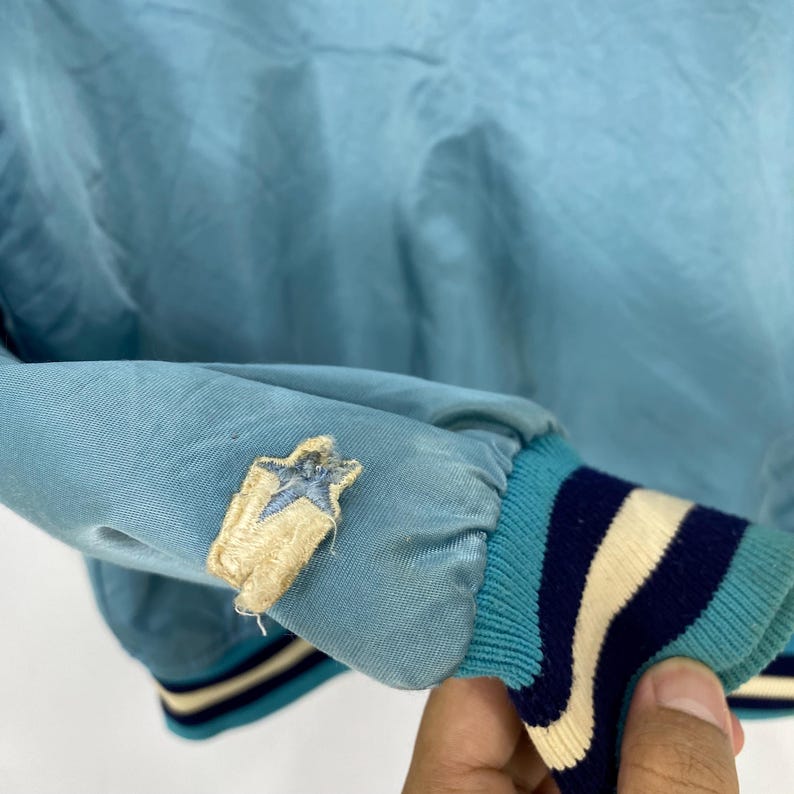 May include: Light blue jacket sleeve with a star patch. The sleeve has a ribbed cuff with stripes in teal, navy, and cream. The star patch is embroidered with a light blue star on a tan background.