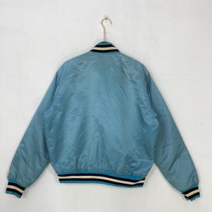 May include: A light blue bomber jacket with a ribbed collar, cuffs, and hem. The jacket has a striped design in navy blue and cream. The jacket is hanging on a gold metal hook.