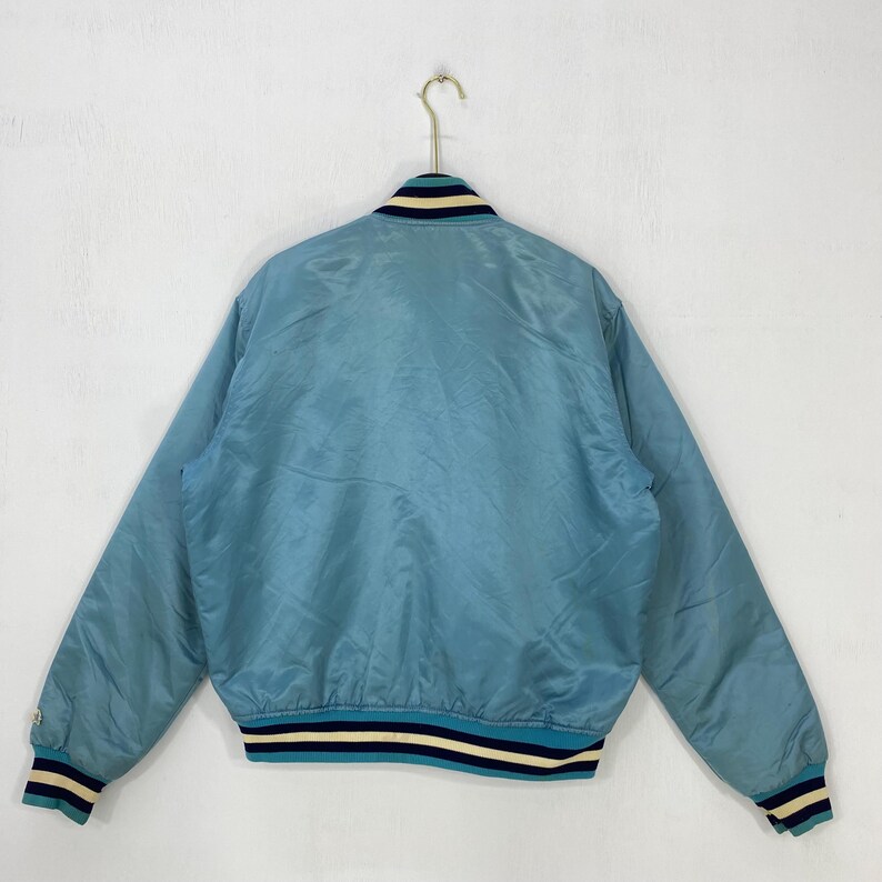 May include: A light blue bomber jacket with a ribbed collar, cuffs, and hem. The jacket has a striped design in navy blue and cream. The jacket is hanging on a gold metal hook.