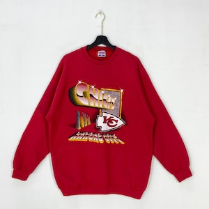 May include: A red long-sleeve crewneck sweatshirt featuring a graphic print on the front. The design includes the word CHIEFS in metallic lettering, the letters NFL, a Kansas City arrowhead logo, and the text KANSAS CITY in a yellow font.