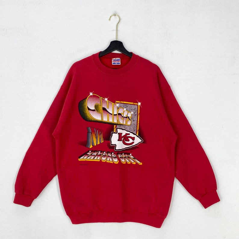 May include: A red long-sleeve crewneck sweatshirt featuring a graphic print on the front. The design includes the word CHIEFS in metallic lettering, the letters NFL, a Kansas City arrowhead logo, and the text KANSAS CITY in a yellow font.