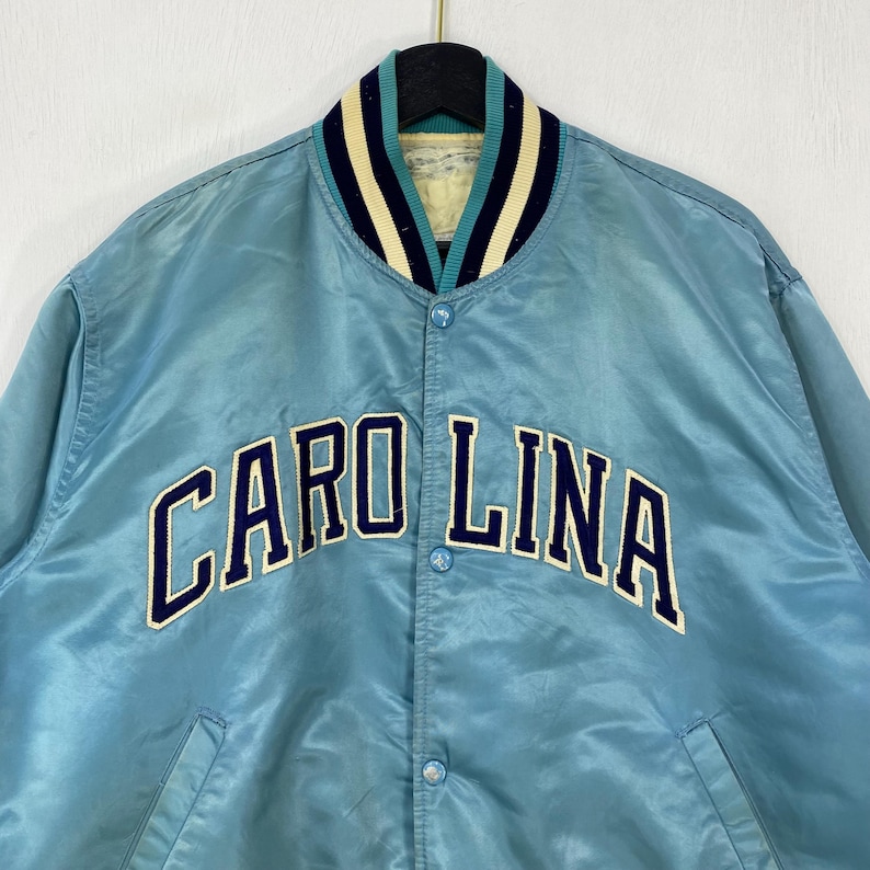 May include: Light blue satin jacket with the word "CAROLINA" in navy blue block letters. The jacket has a striped collar in navy, white, and teal. The jacket has a button-up front and two pockets.