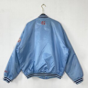 Puede incluir: Chaqueta bomber de sat&eacute;n azul claro con cuello, pu&ntilde;os y dobladillo de canal&eacute;. Presenta un logotipo rojo y blanco en la espalda y la manga. La chaqueta est&aacute; colgada de una percha met&aacute;lica contra una pared blanca.