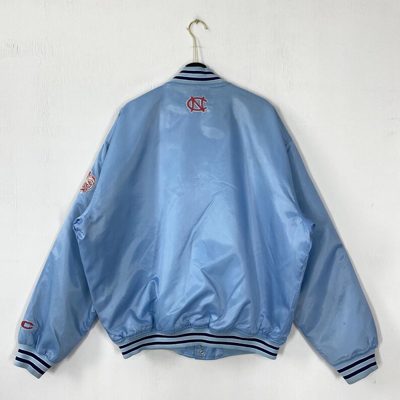 Puede incluir: Chaqueta bomber de sat&eacute;n azul claro con cuello, pu&ntilde;os y dobladillo de canal&eacute;. Presenta un logotipo rojo y blanco en la espalda y la manga. La chaqueta est&aacute; colgada de una percha met&aacute;lica contra una pared blanca.