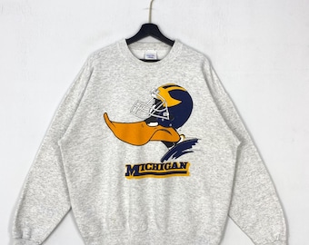 May include: A light grey crewneck sweatshirt featuring a graphic of a duck wearing a dark blue and yellow football helmet. Below the duck, the word MICHIGAN is printed in dark blue letters with a yellow outline. The sweatshirt has long sleeves.