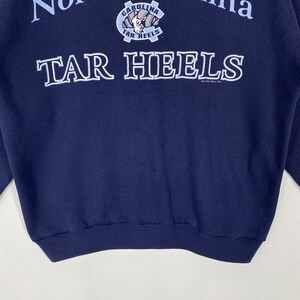 May include: Navy blue sweatshirt featuring the text "TAR HEELS" in white, bold lettering. Above the text is a logo with the words "CAROLINA" and "TAR HEELS". The sweatshirt is a solid navy blue colour.