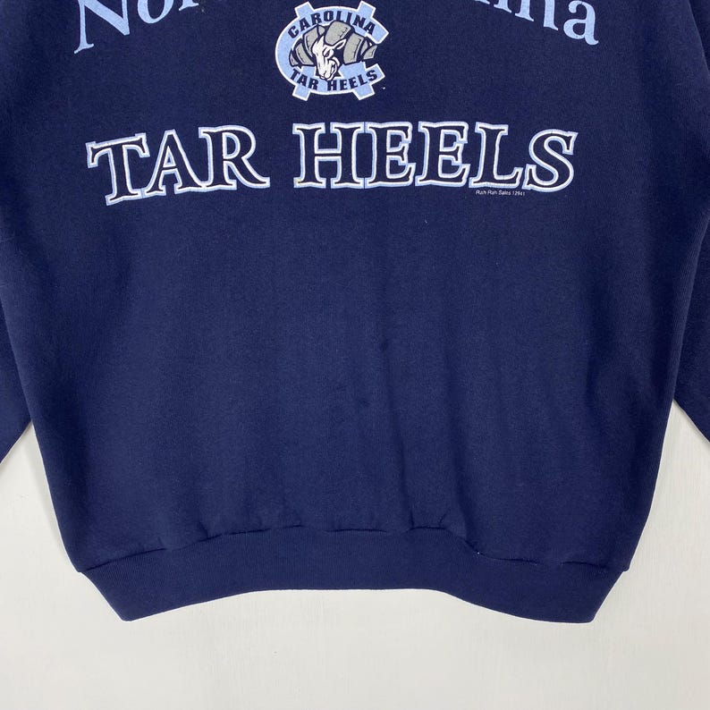 May include: Navy blue sweatshirt featuring the text "TAR HEELS" in white, bold lettering. Above the text is a logo with the words "CAROLINA" and "TAR HEELS". The sweatshirt is a solid navy blue colour.