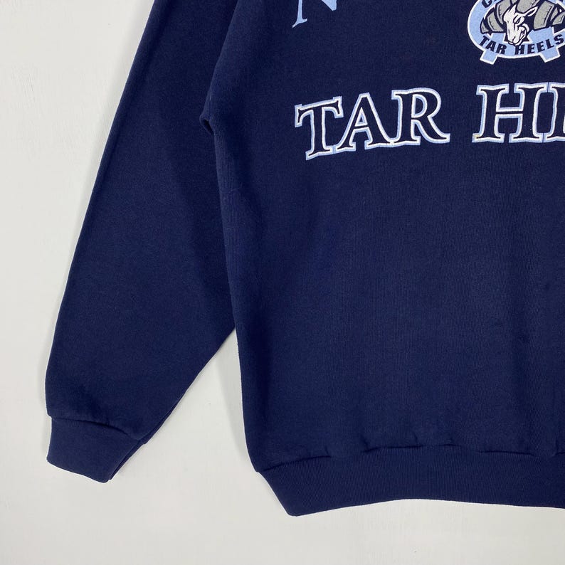 May include: Navy blue sweatshirt with the text "TAR HEELS" in white block letters. The sweatshirt also features a light blue emblem with the words "TAR HEELS" and a ram graphic. The sleeve and bottom hem are ribbed.
