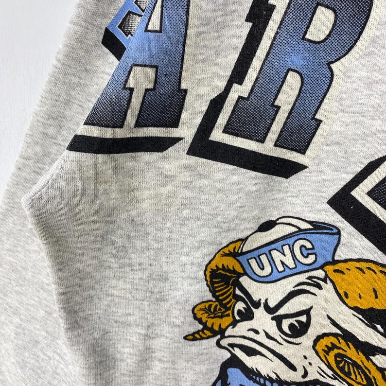 May include: A light grey heathered sweatshirt featuring a large graphic print. The design includes bold, dark blue block lettering and a cartoon ram mascot wearing a light blue sailor hat with the white text UNC printed on the front.