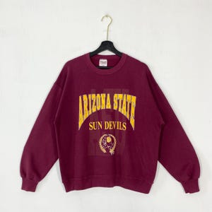 Op de afbeelding: Bordeauxkleurige Arizona State Sun Devils sweatshirt met lange mouwen. De voorkant heeft de woorden "ARIZONA STATE" in gele gebogen letters boven "SUN DEVILS". Een cirkelvormig embleem staat eronder.