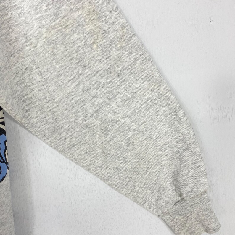 May include: A light grey heathered long-sleeve sweatshirt sleeve featuring a ribbed cuff at the wrist. The fabric has a speckled texture throughout, and a portion of a graphic design with blue and black elements is visible along the left side.