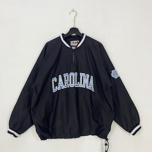 May include: Black pullover jacket with "CAROLINA" in light blue lettering across the chest. The jacket has a half-zip closure, a drawstring hem, and a small logo patch on the sleeve. White and blue striped trim accents the collar and cuffs.