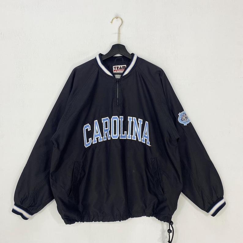 May include: Black pullover jacket with "CAROLINA" in light blue lettering across the chest. The jacket has a half-zip closure, a drawstring hem, and a small logo patch on the sleeve. White and blue striped trim accents the collar and cuffs.