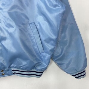 Puede incluir: Chaqueta de sat&eacute;n azul claro con un bolsillo en la parte delantera. La chaqueta tiene un dobladillo y pu&ntilde;os acanalados con rayas azul marino y azul claro. La tela tiene una textura brillante y suave.