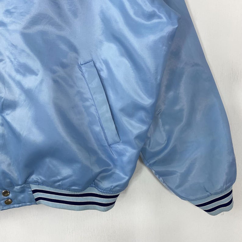 Puede incluir: Chaqueta de sat&eacute;n azul claro con un bolsillo en la parte delantera. La chaqueta tiene un dobladillo y pu&ntilde;os acanalados con rayas azul marino y azul claro. La tela tiene una textura brillante y suave.