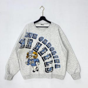 May include: A heather grey crewneck sweatshirt featuring the text NORTH CAROLINA TAR HEELS in blue and black block lettering. A graphic of a ram mascot wearing a blue sweater with the letters UNC is positioned on the lower left side.