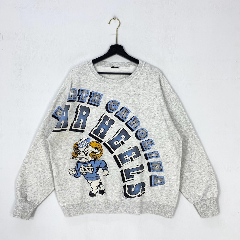 May include: A heather grey crewneck sweatshirt featuring the text NORTH CAROLINA TAR HEELS in blue and black block lettering. A graphic of a ram mascot wearing a blue sweater with the letters UNC is positioned on the lower left side.