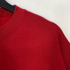 May include: A solid red crewneck sweatshirt featuring a ribbed collar and a soft fleece interior lining. The garment is displayed on a black hanger, showing a plain, unpatterned fabric texture across the shoulder and chest area.