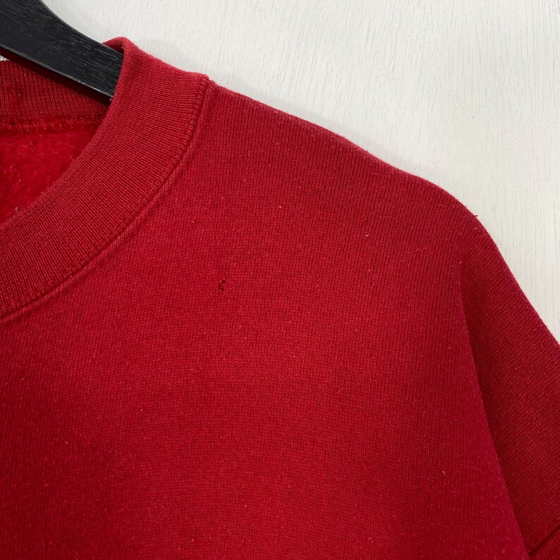 May include: A solid red crewneck sweatshirt featuring a ribbed collar and a soft fleece interior lining. The garment is displayed on a black hanger, showing a plain, unpatterned fabric texture across the shoulder and chest area.