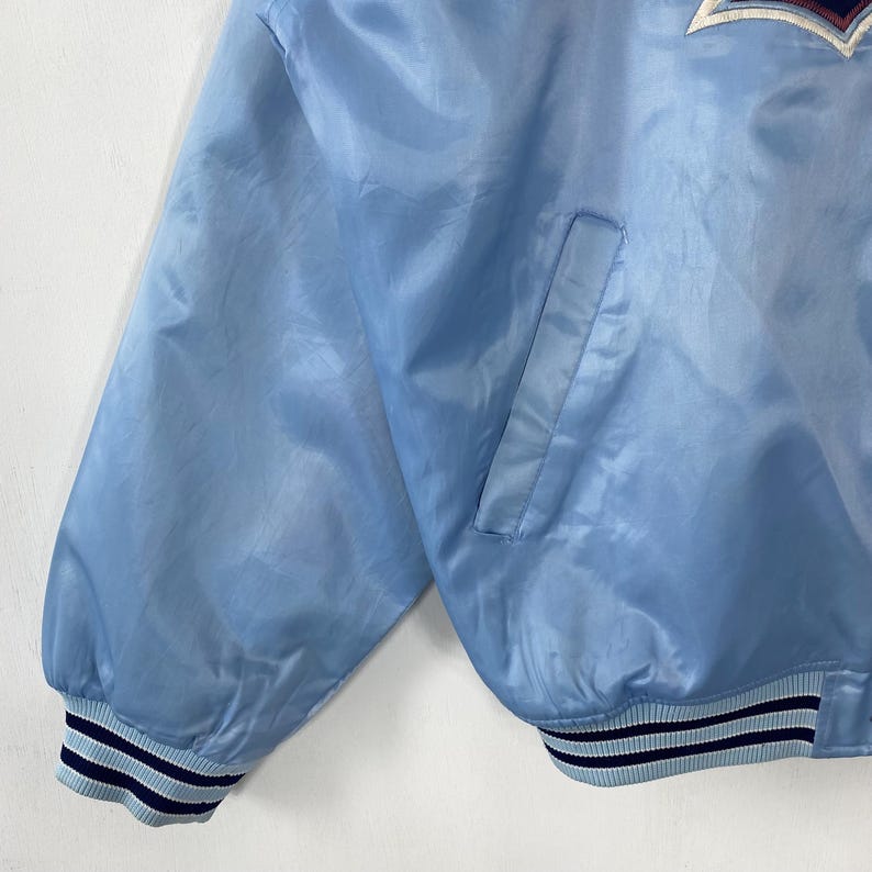 Puede incluir: Chaqueta bomber de sat&eacute;n azul claro con bolsillo y pu&ntilde;os acanalados. Los pu&ntilde;os tienen rayas azul marino y blancas. La chaqueta tiene un logotipo en la parte superior del pecho.
