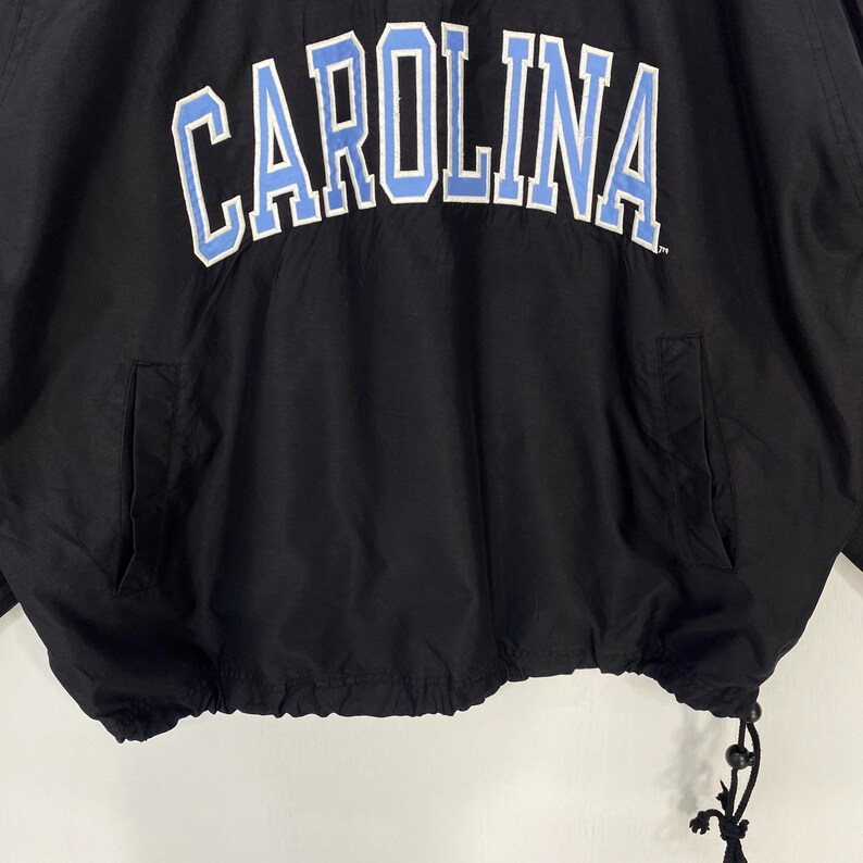 May include: Black jacket with the word "CAROLINA" in white letters with a light blue outline across the chest. The jacket has two front pockets and a drawstring at the bottom. The jacket is made of a lightweight material.