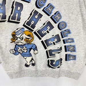 May include: A heather grey sweatshirt featuring a graphic of a ram mascot wearing a blue sweater with the letters NC and a sailor hat labelled UNC. The text NORTH CAROLINA TAR HEELS is printed in large, arched, blue and black block lettering.