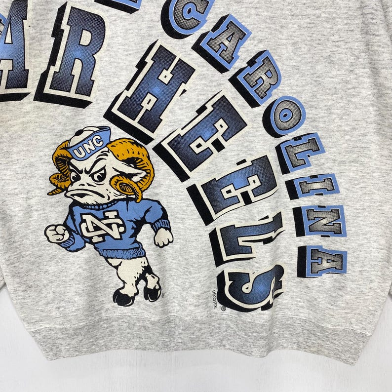 May include: A heather grey sweatshirt featuring a graphic of a ram mascot wearing a blue sweater with the letters NC and a sailor hat labelled UNC. The text NORTH CAROLINA TAR HEELS is printed in large, arched, blue and black block lettering.