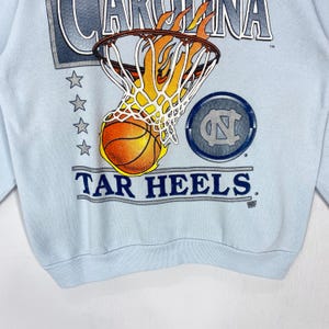 May include: A light blue sweatshirt featuring a graphic of a basketball passing through a flaming hoop. The text reads CAROLINA and TAR HEELS, accompanied by a circular logo with the letters NC and a vertical row of four grey stars on the left side.
