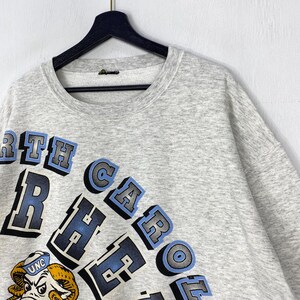 May include: A light grey heathered crewneck sweatshirt featuring the text NORTH CAROLINA TAR HEELS in a bold, arched blue and black font. Below the text is a graphic illustration of a ram mascot wearing a blue hat with the letters UNC.