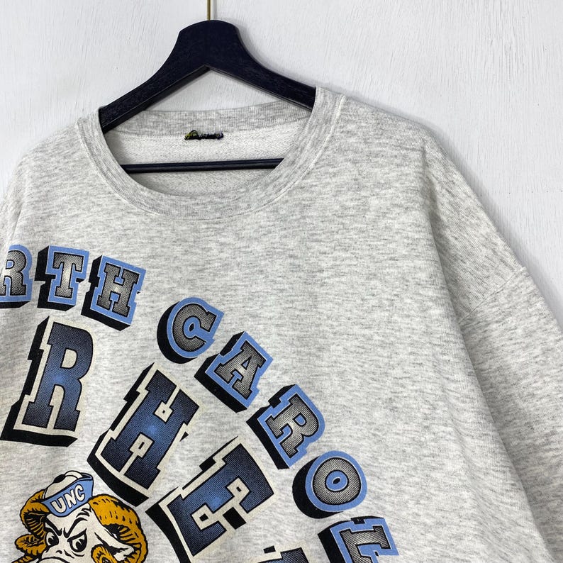 May include: A light grey heathered crewneck sweatshirt featuring the text NORTH CAROLINA TAR HEELS in a bold, arched blue and black font. Below the text is a graphic illustration of a ram mascot wearing a blue hat with the letters UNC.