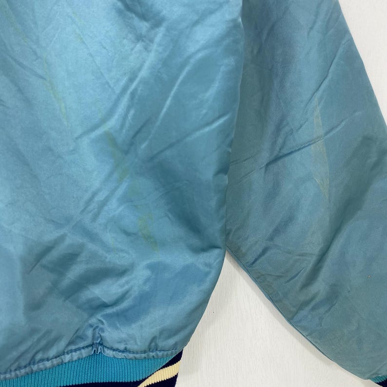 May include: A light blue satin jacket with a ribbed collar and cuffs. The jacket has a quilted design and a slight sheen. The cuffs are striped with blue, white, and navy. The jacket appears to be vintage.