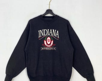 May include: A black long-sleeve crewneck sweatshirt featuring an embroidered Indiana Hoosiers logo on the chest. The text reads INDIANA, EST. 1820, HOOSIERS, and BLOOMINGTON, IN. The design includes a red and white emblem with green accents.