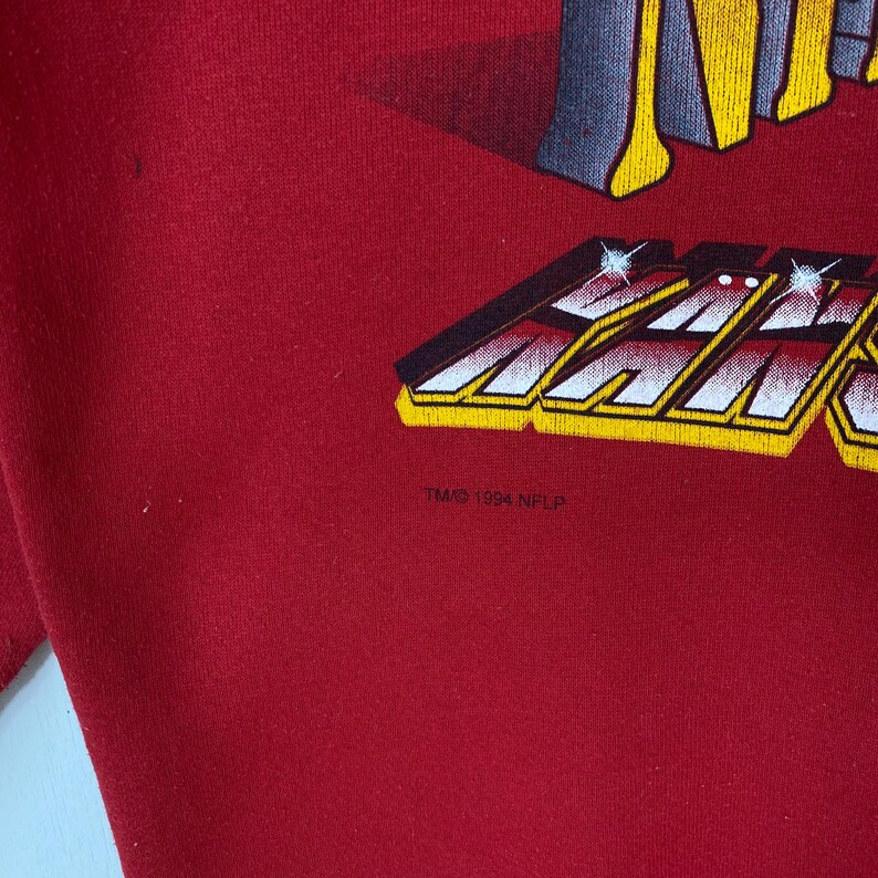 May include: A red sweatshirt featuring a graphic print with yellow and white lettering. The design includes the text "KANSAS" in a stylized font with a metallic effect and sparkles. Below the graphic, the text "TM/&copy; 1994 NFLP" is printed.