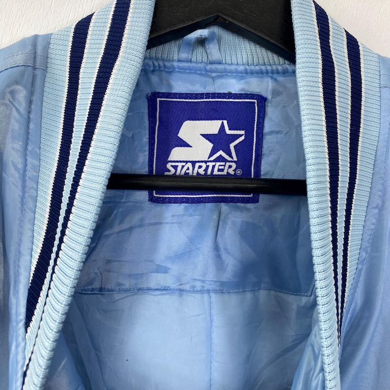Puede incluir: Chaqueta Starter azul claro con cuello y ribetes a rayas azul marino y blanco. La chaqueta presenta un parche con el logo de Starter en el pecho. La chaqueta es de un material brillante, posiblemente nailon.